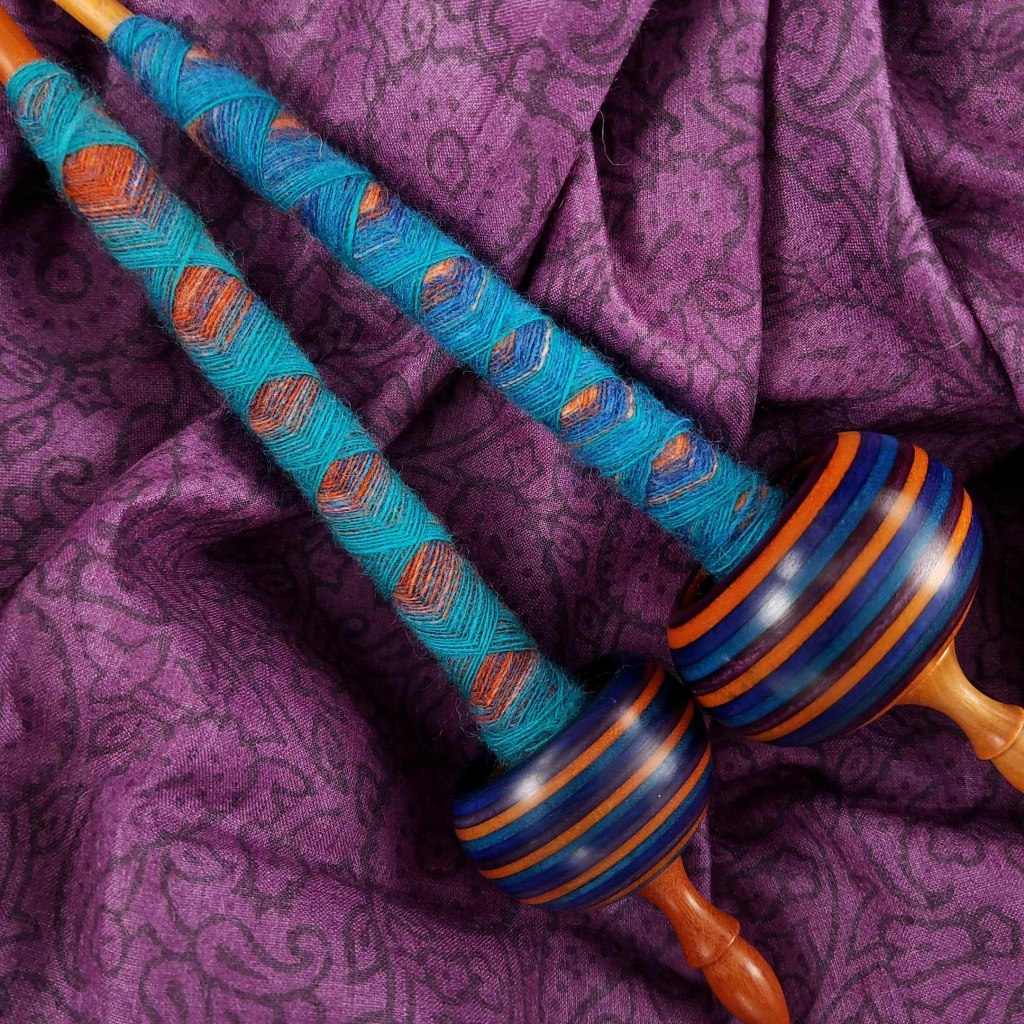 Two supported spindles in orange, blue, turquoise and purple both have fancily wrapped cops of fiber in the same colors