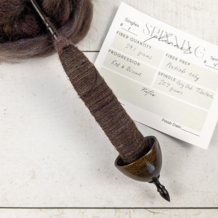 A bog oak Tibetan spindle with some spinning on it, and a spinning planner card and more fiber behind it