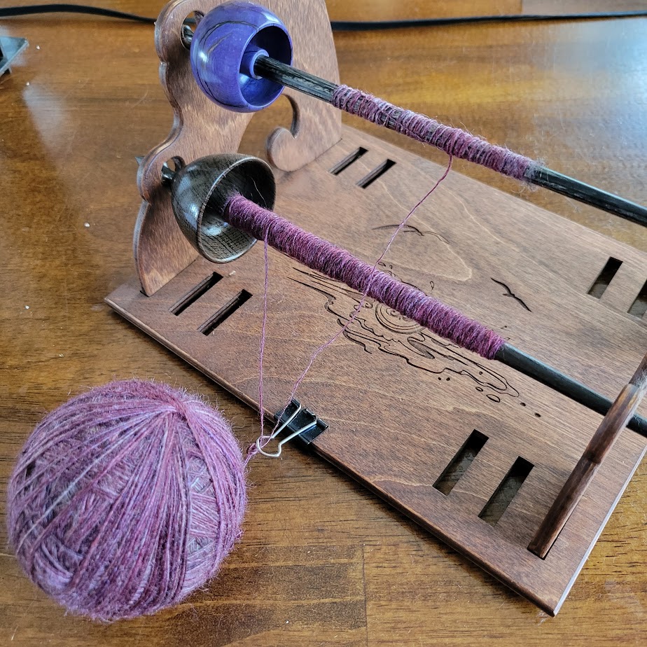Two spindles in a plying stand, with the singles passing through a binder clip on the stand before getting wrapped into a large ball
