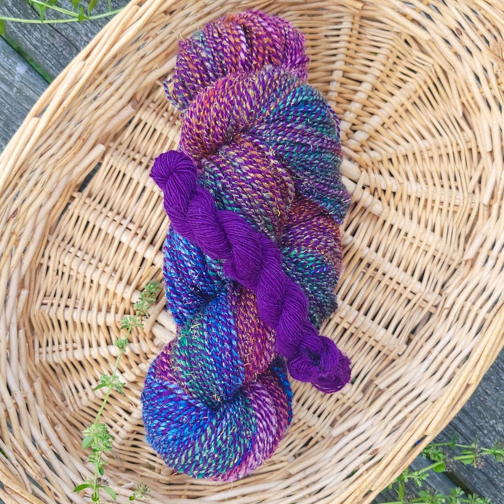 Two skeins of yarn - one barberpoled two-ply in purples, blues and pinks, and one singles yarn in purple - sit in a basket
