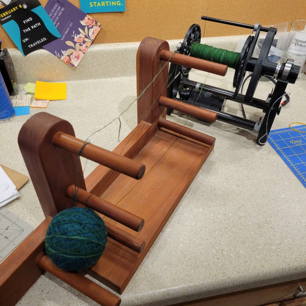 An inkle loom sits on a counter between a bobbin holding singles yarn, and partially wrapped chain plying ball.