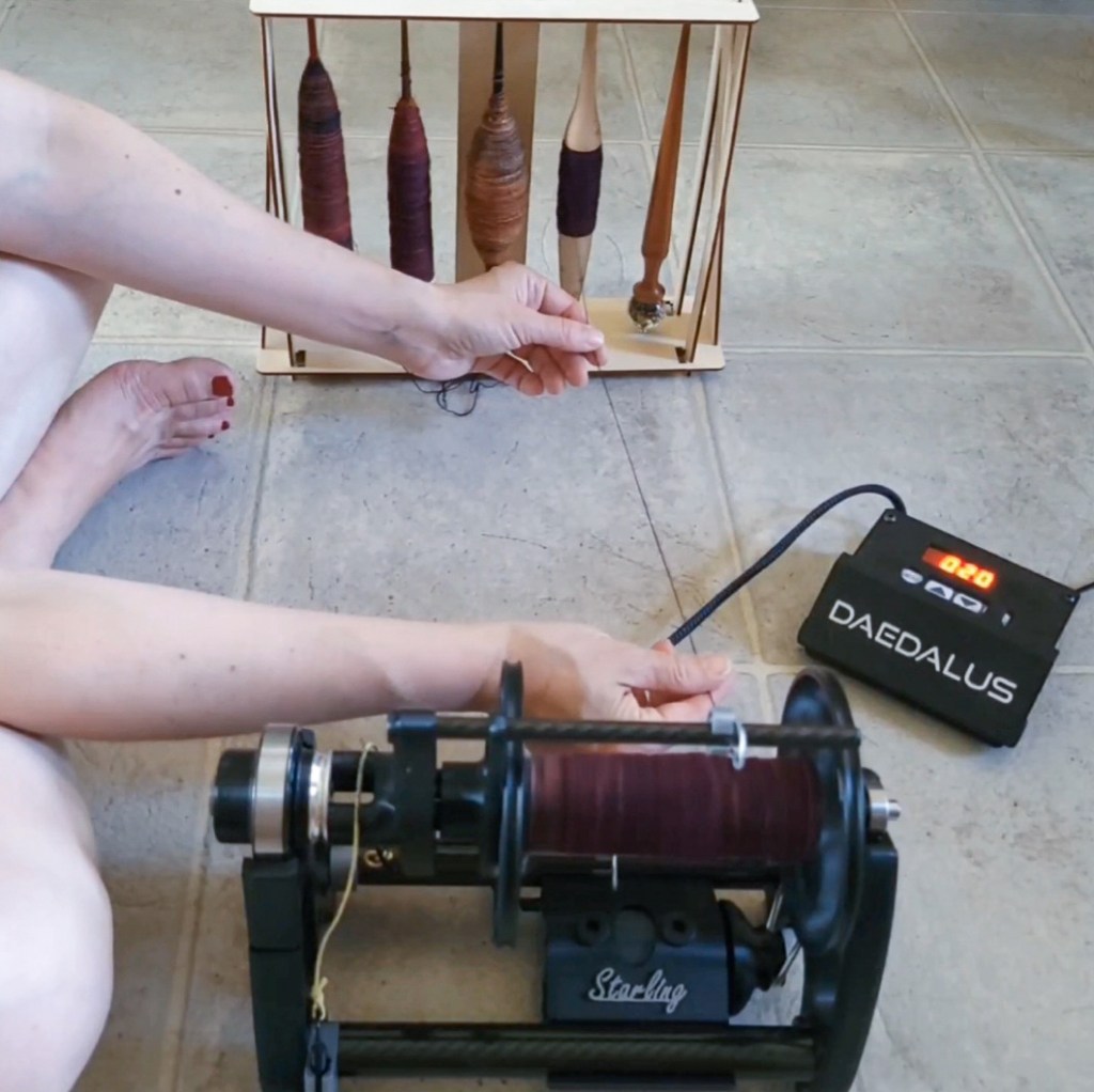 An eSpinner being used to reel singles off five spindles in a storage stand, with a person sitting on the kitchen floor making sure the singles feed smoothly