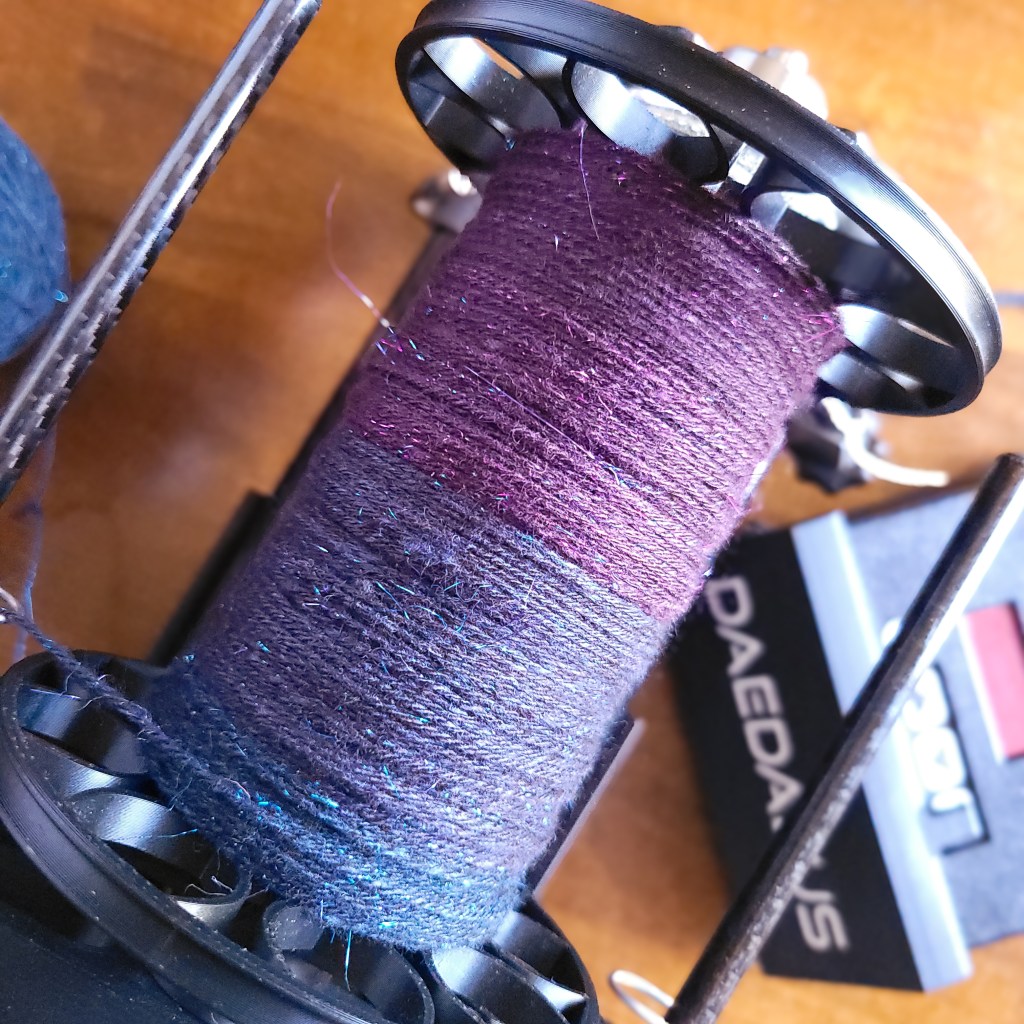 An overhead shot of a Daedalus Starling, partway through adding twist to a yarn in various sparkly shades of blue and purple.
