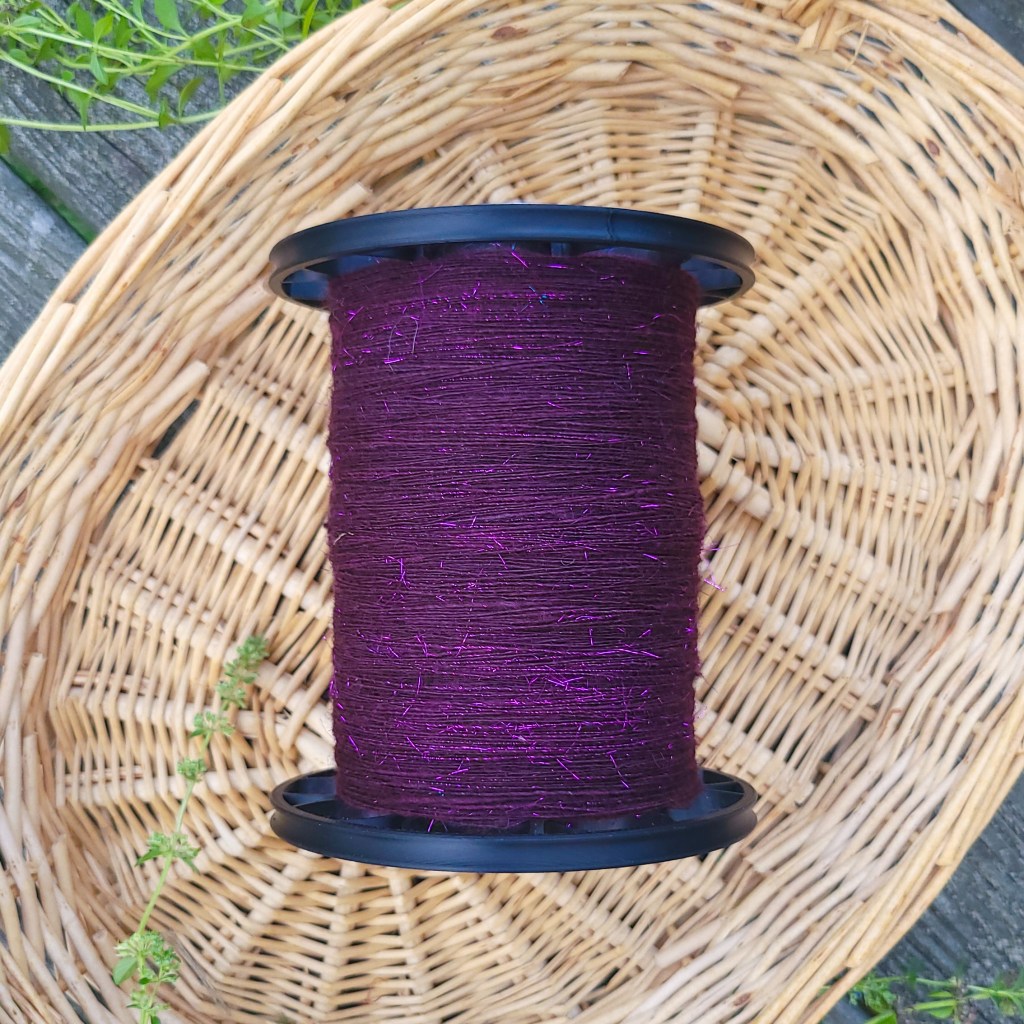 A bobbin of sparkly purple singles yarn rests in a basket