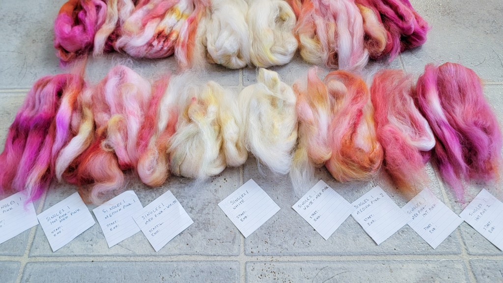 A row of pink and white fiber, representing one ply of the three-ply yarn, is laid out on the floor with paper labels identifying each chunk of fiber.
