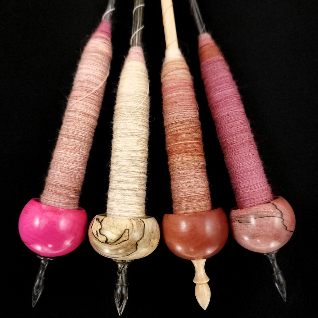 Four ninja-style supported spindles rest on a black background. The spindles have moderately-sized cops that form the first ply of Tickled Pink.