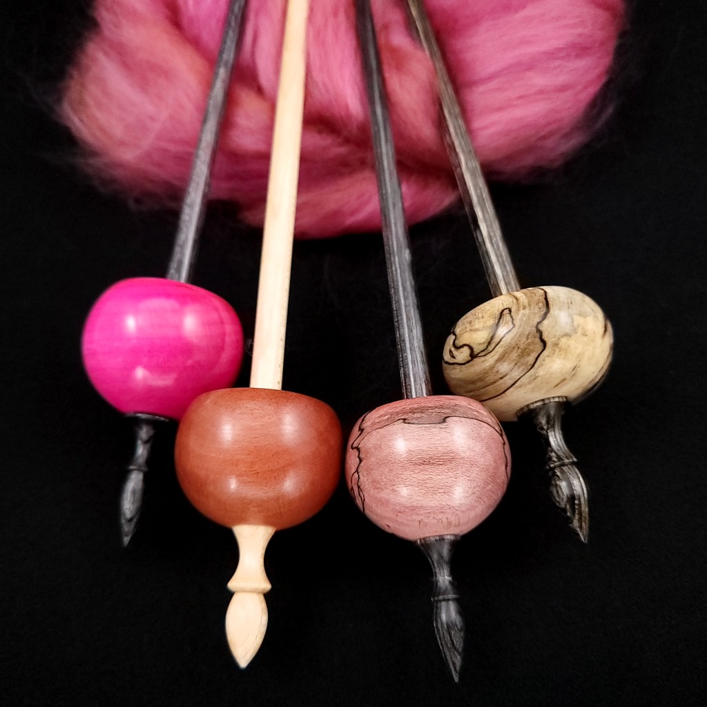Four ninja-style supported spindles rest on a black background with their tips nestled in a bundle of hot pink fiber