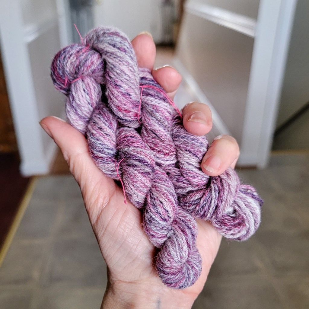 A hand holds a skein of chain-plied pink, purple and white yarn