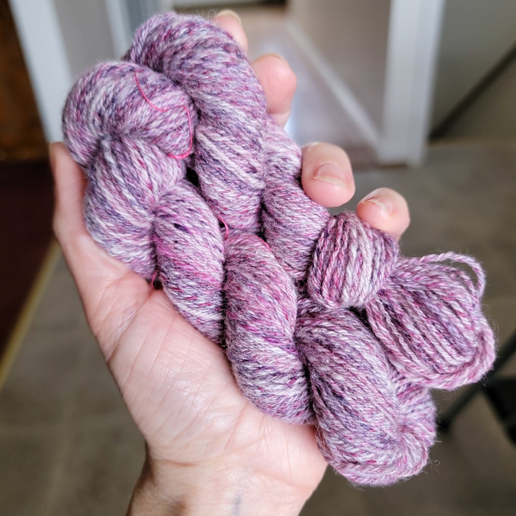 A hand holds a skein of three-ply yarn of pink, purple and white