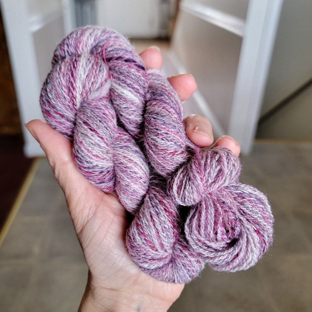 A hand holds a skein of two-ply yarn of pink, purple and white