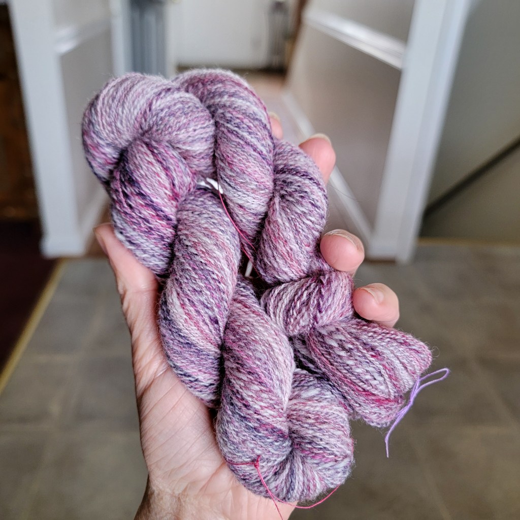 A hand holds a skein of two-ply yarn of pink, purple and white