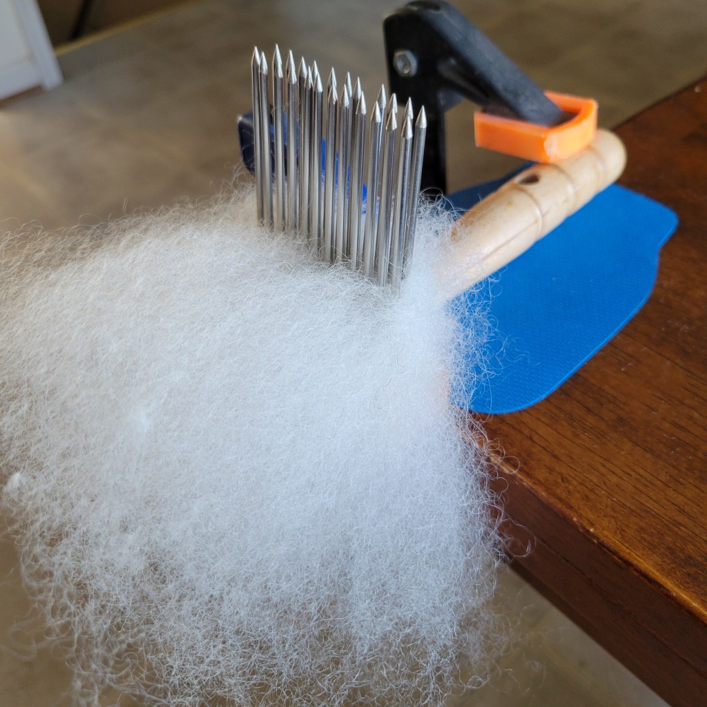 A wool comb with a blue nonslip mat is clamped to a brown table. Partially combed white wool is in the teeth of the comb.